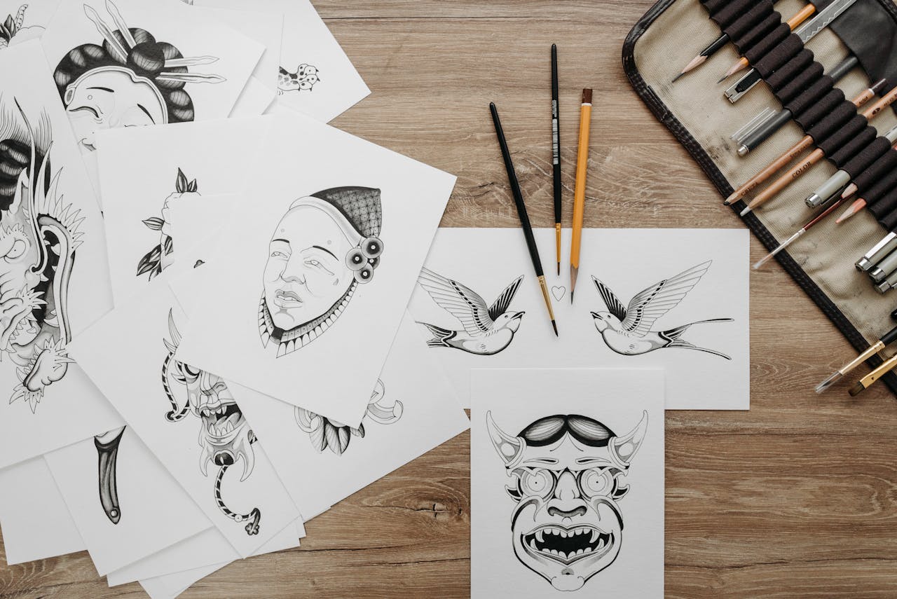 Flat lay of intricate tattoo sketches and drawing tools on wooden table.